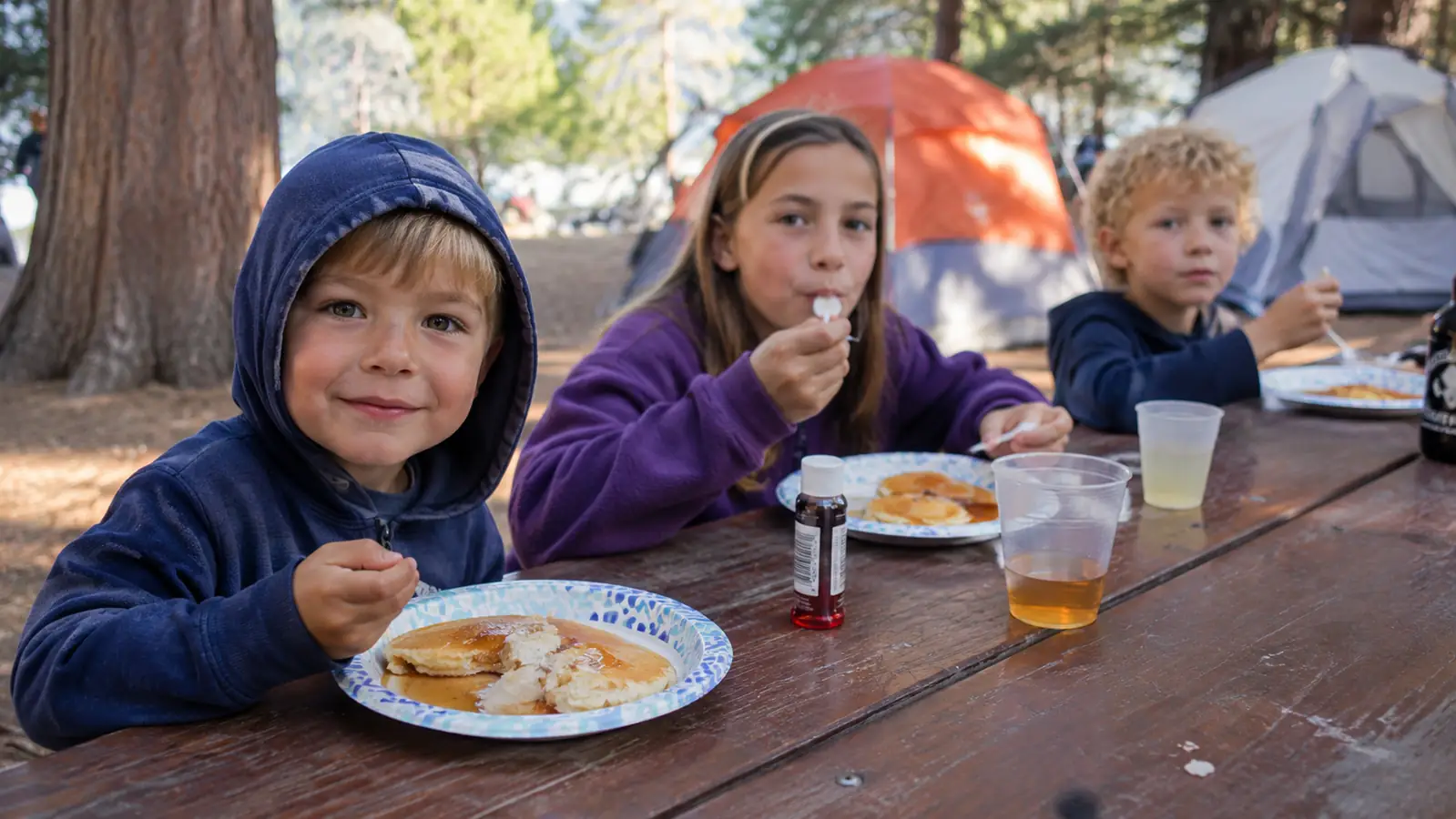 A group of kids eating pancakes at a long picnic table at a campsite